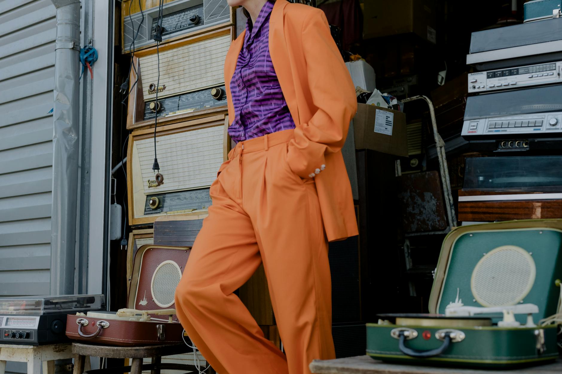 A fashionable person in an orange suit leaning against retro stereos in a store.