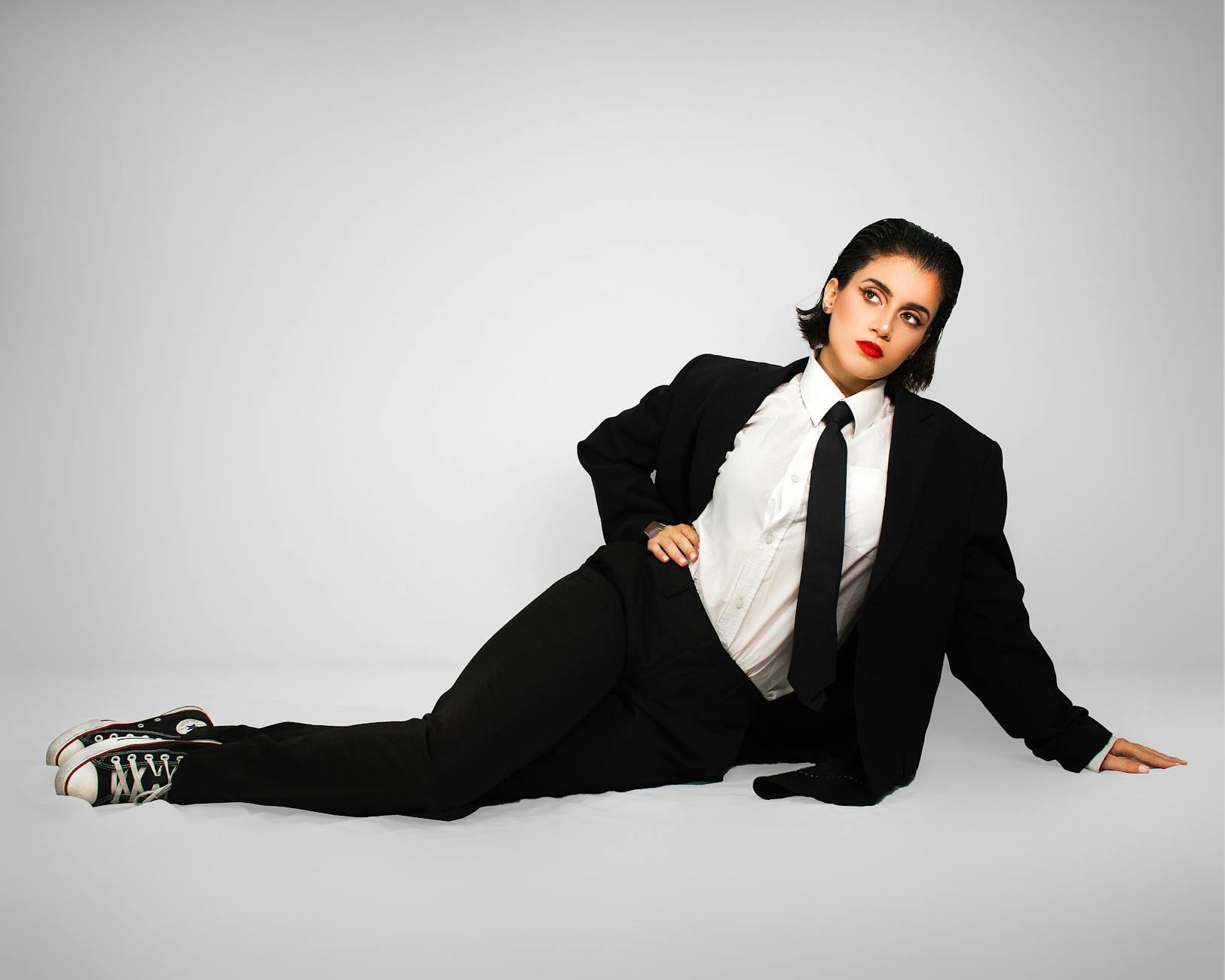 Confident woman in black suit and tie reclining in studio for a modern fashion portrait.
