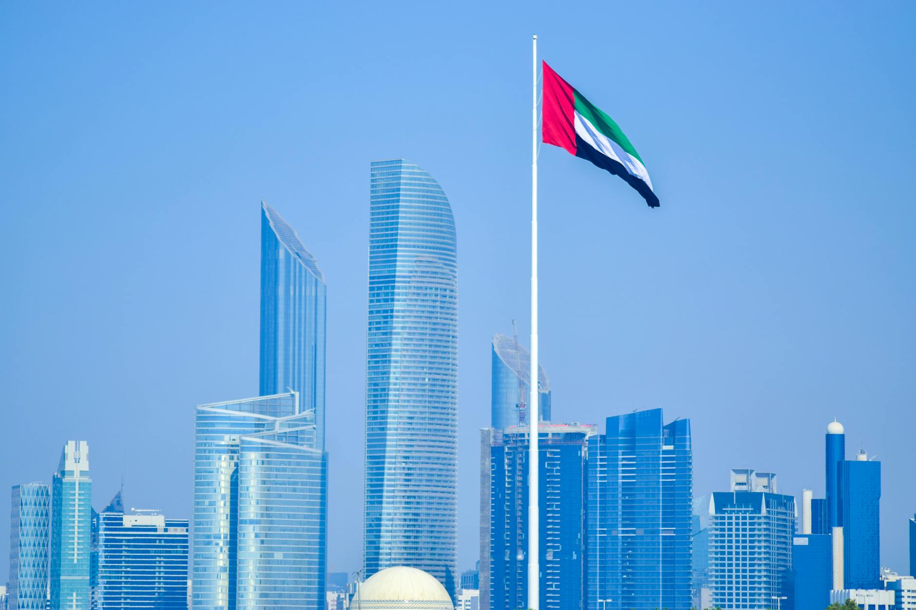 Modern skyline of UAE cityscape with national flag waving against clear sky.
