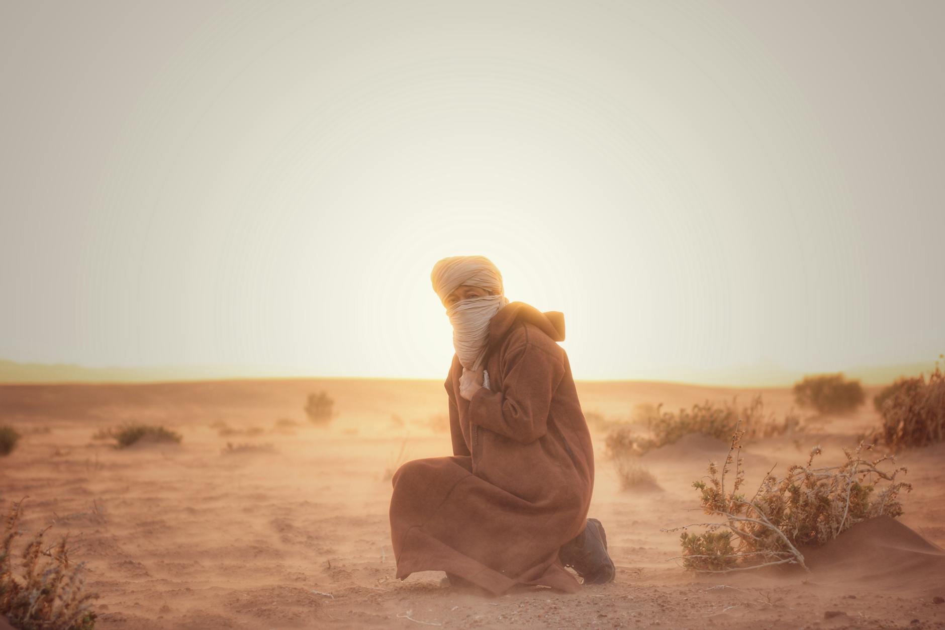 A man in traditional Moroccan attire kneels in an arid desert at sunset.