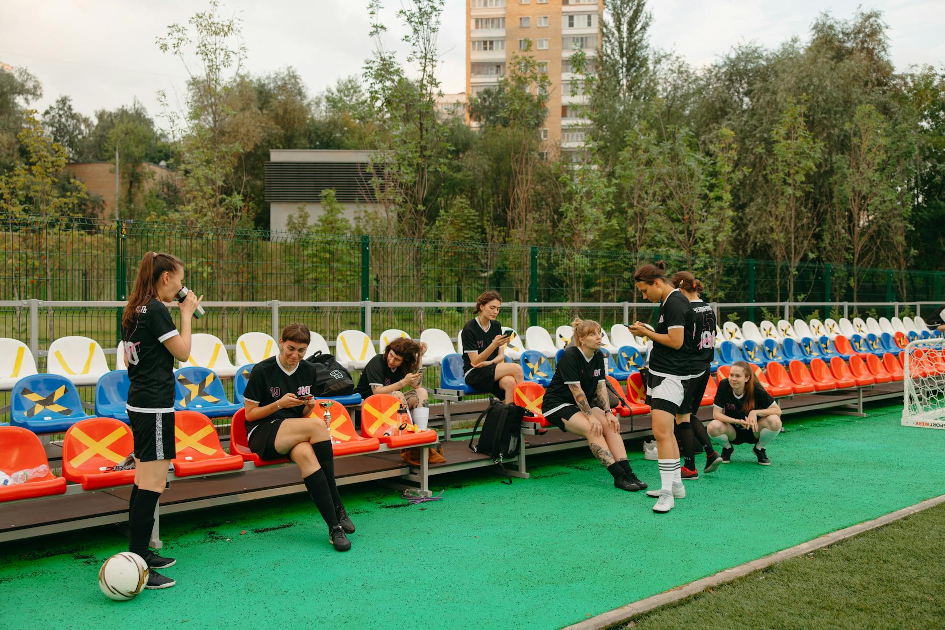 Women's football team relaxing on a sports field, sitting on benches and interacting with each other.